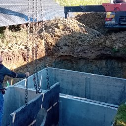 Firma-handlowo-usługow transportowa,,PE-TOM ''Krzysztof Pestka - Montaż betonowego zbiornika w wykopie, z użyciem dźwigu. Widoczne panele słoneczne w tle. Praca ziemna w toku, teren nierówny.