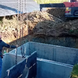 Montaż betonowego zbiornika w wykopie, z użyciem dźwigu. Widoczne panele słoneczne w tle. Praca ziemna w toku, teren nierówny.