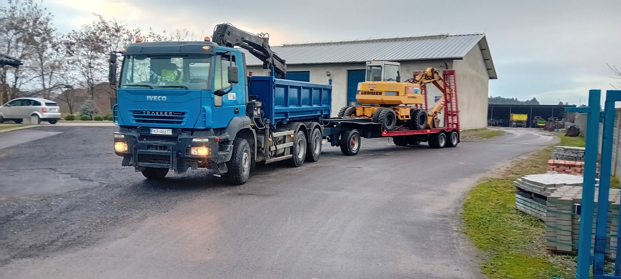 Niebieska ciężarówka Iveco z HDS przewozi żółtą koparkę Liebherr na czerwonej platformie. W tle budynek gospodarczy i zaparkowany samochód osobowy.