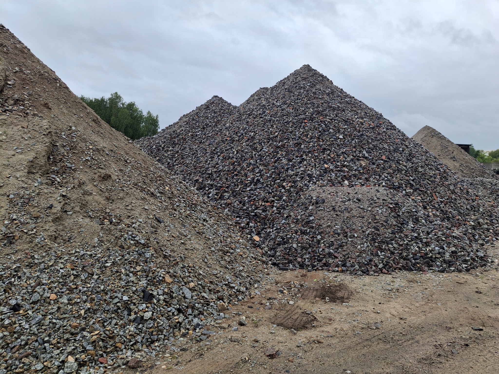 Góra piasku i gruzu pod pochmurnym niebem. Widoczne różne frakcje materiału. Na horyzoncie zieleń drzew. Składowisko materiałów budowlanych.