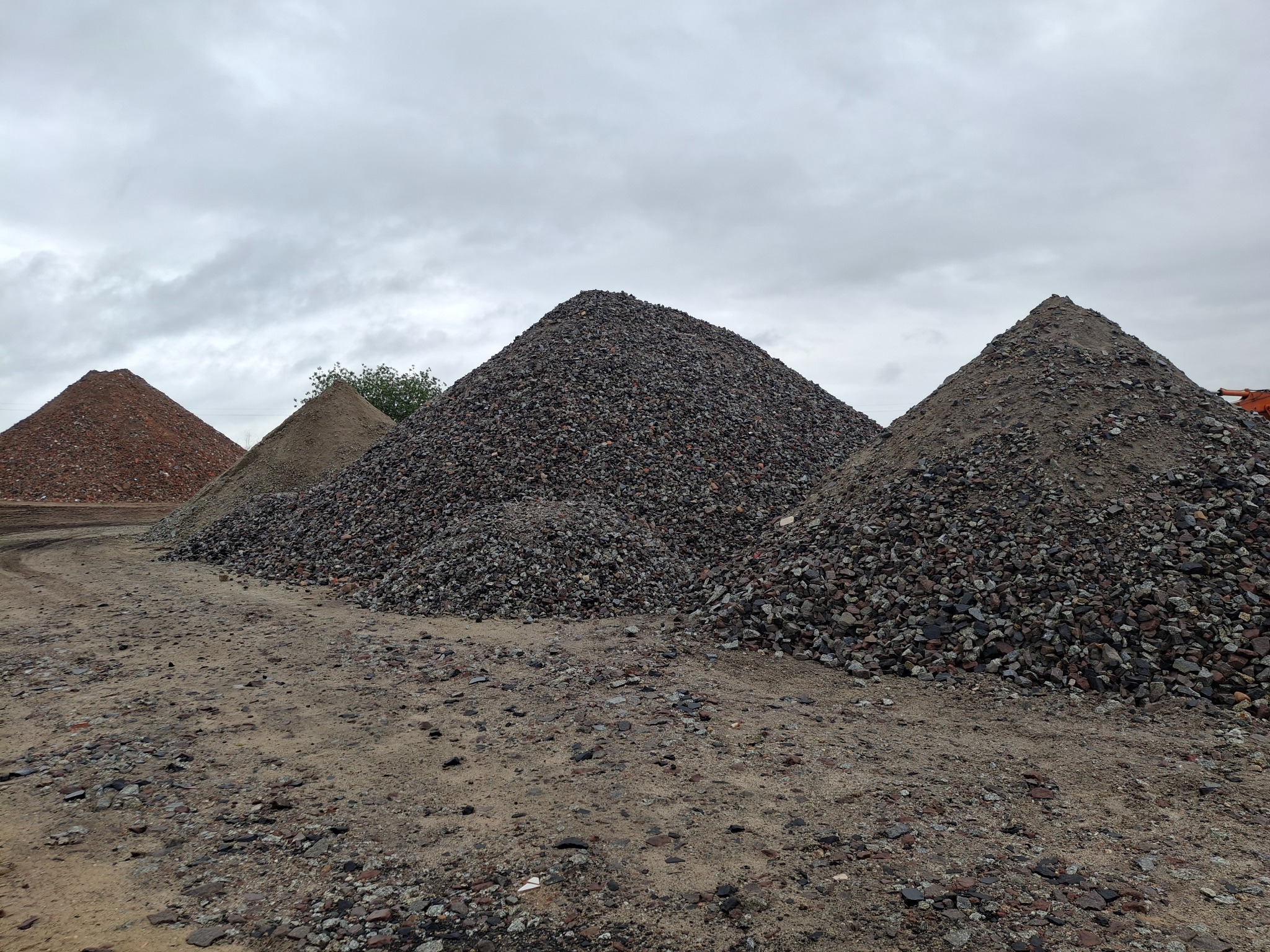 Góra ziemi, piasku i gruzu o różnej frakcji pod pochmurnym niebem. Na horyzoncie widoczny pomarańczowy element maszyny budowlanej.