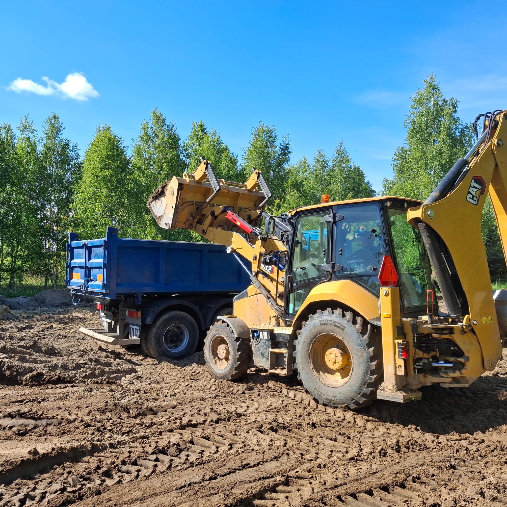 Żółta koparko-ładowarka CAT nabiera ziemię i wsypuje ją do niebieskiej ciężarówki na tle zielonych drzew i błękitnego nieba. Praca w terenie.