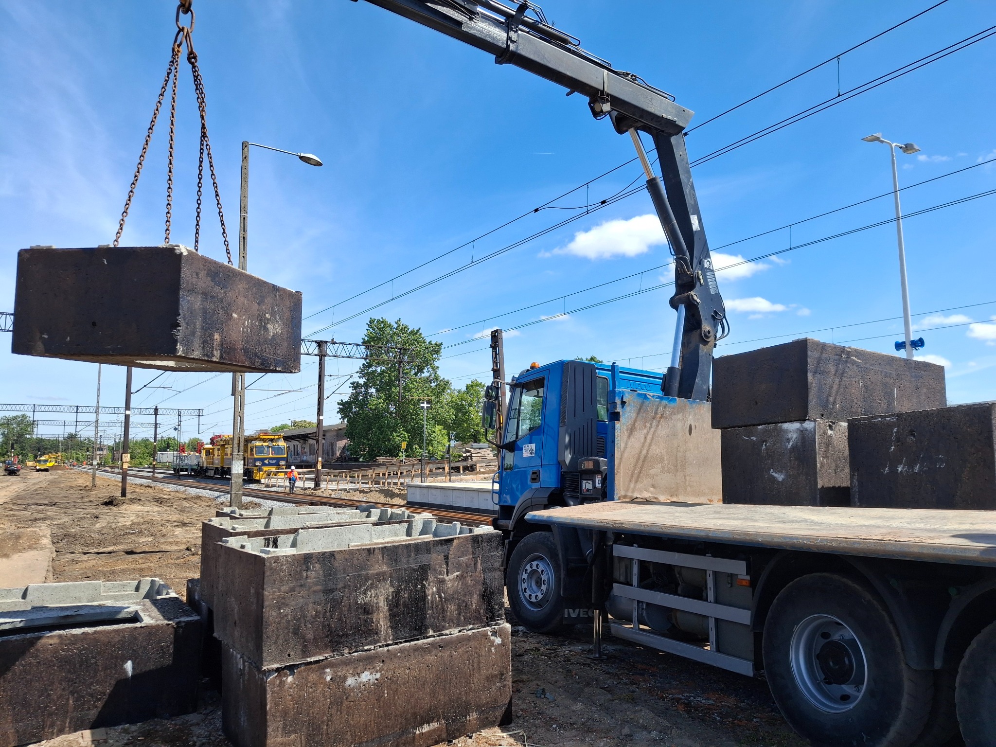 Żuraw samochodowy podnosi betonowy element na tle torów kolejowych i błękitnego nieba. Widoczne elementy infrastruktury kolejowej i inne maszyny.