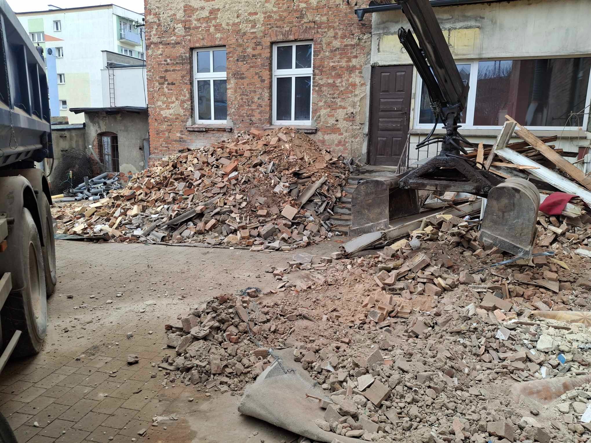 Sterta gruzu ceglanego przed budynkiem z cegły, widoczny fragment ciężarówki i ramię dźwigu, teren po rozbiórce.