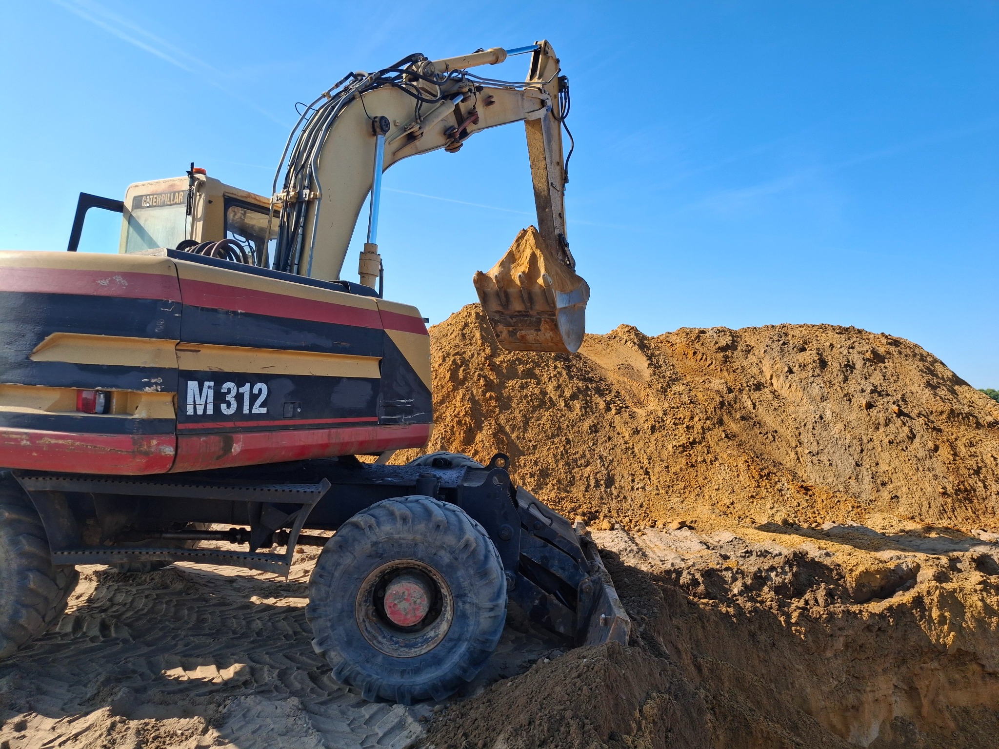 Żółta koparka kołowa Caterpillar M312 nabiera piasek z hałdy na tle błękitnego nieba. Widoczne ślady opon na ziemi.