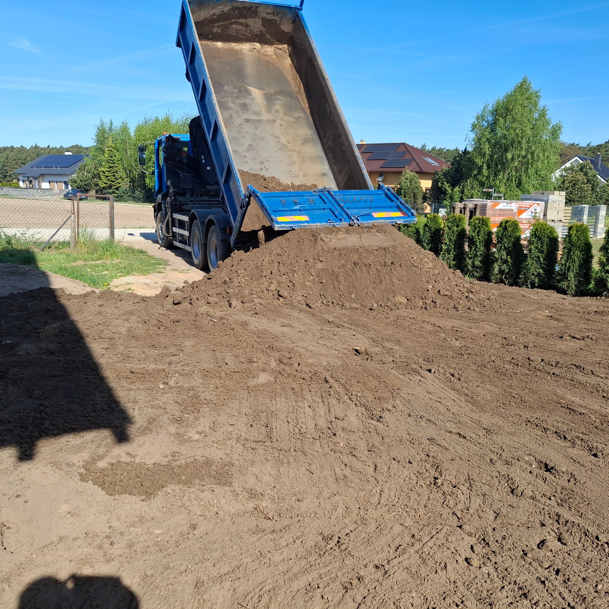 Ciężarówka z uniesioną skrzynią wyładowuje ziemię na placu budowy, widoczne budynki w tle, słoneczny dzień.