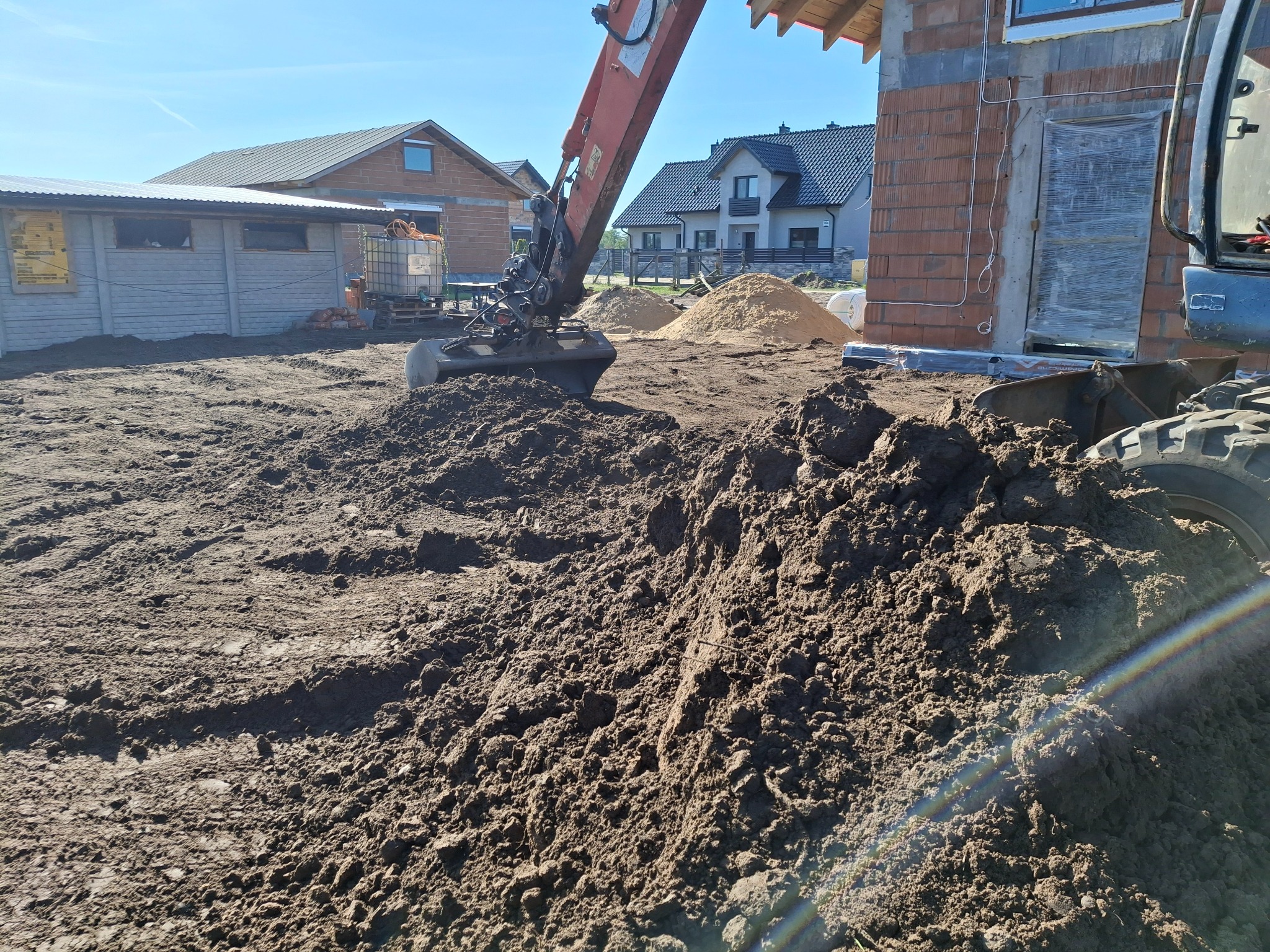 Koparka nabiera ziemię na budowie domu jednorodzinnego. W tle sterta piasku i inne budynki. Widoczne ślady opon na ziemi.