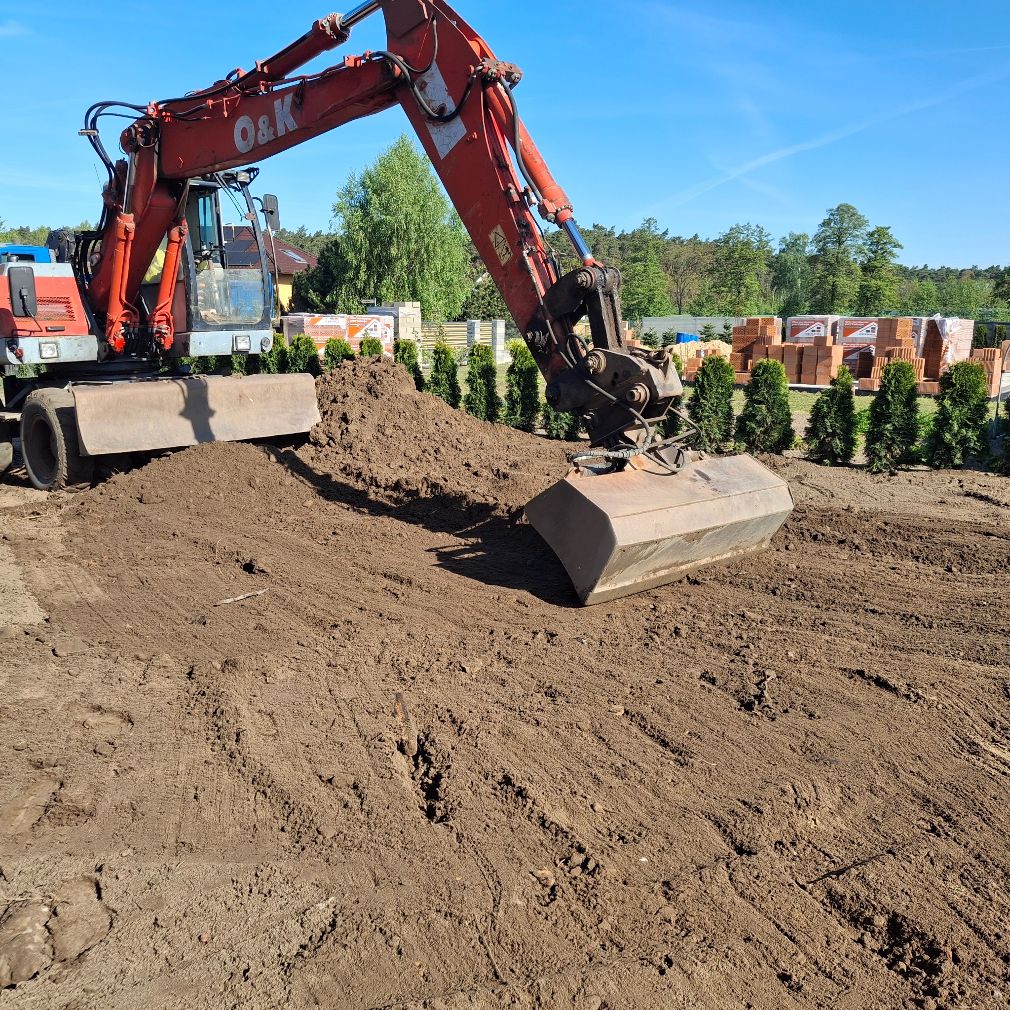 Czerwona koparka O&K wyrównuje teren pod budowę, w tle cegły i tuje, błękitne niebo. Widoczne ślady pracy maszyny na ziemi.