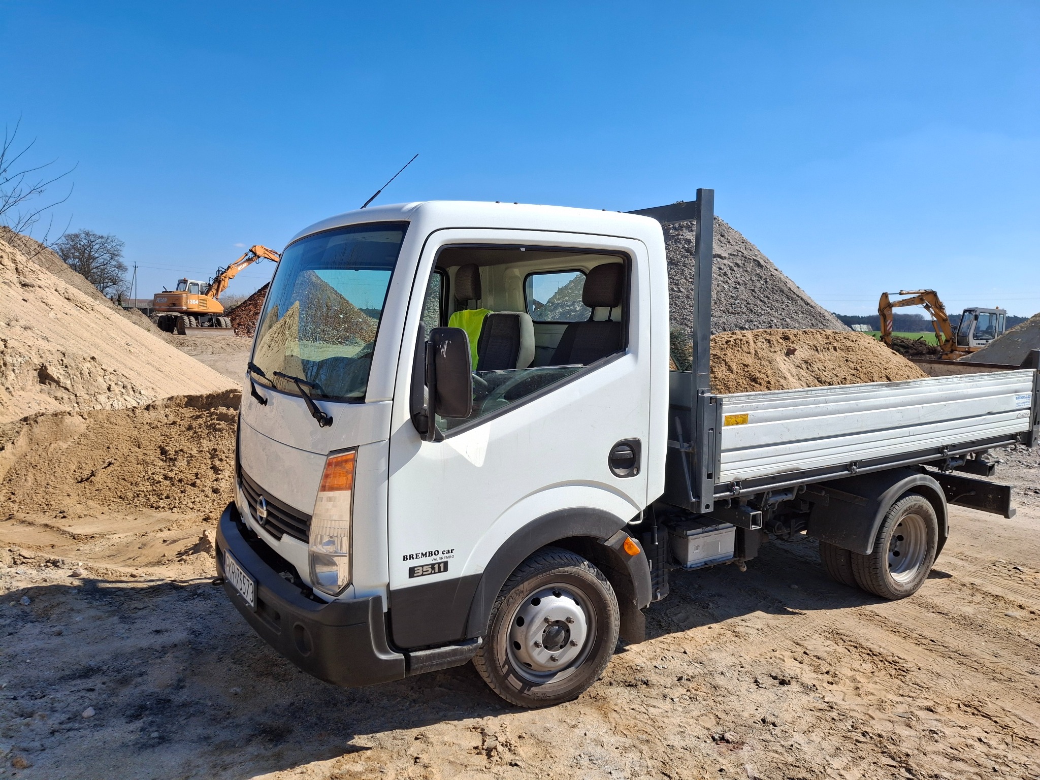 Biała ciężarówka z ładunkiem piasku na pace, w tle koparki i hałdy ziemi. Ujęcie z boku, w słoneczny dzień.