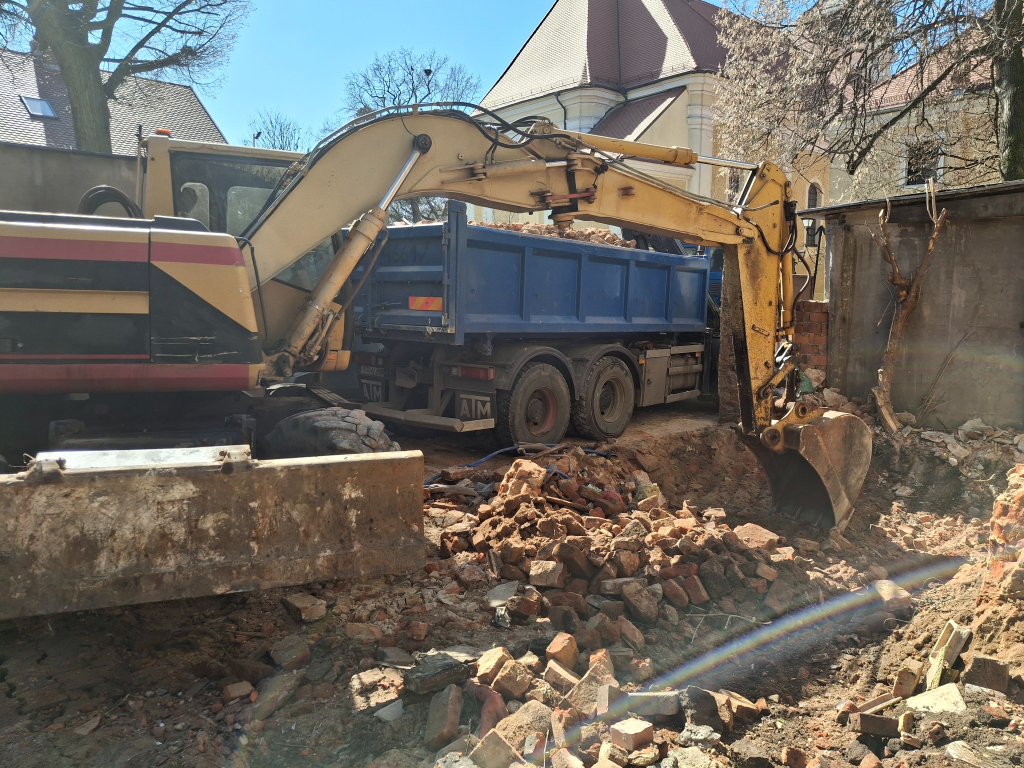 Żółta koparka załadowuje gruz ceglany do niebieskiej ciężarówki na tle budynku. Widoczny lemiesz koparki, fragment ściany i drzewa.