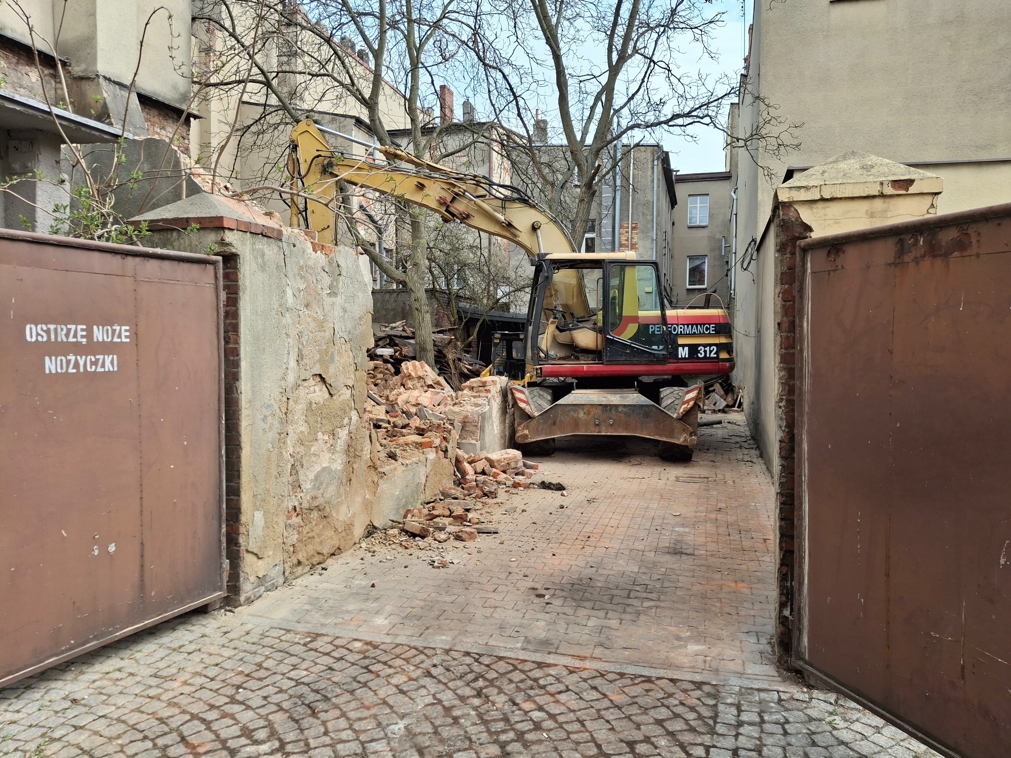Żółta koparka Performance M 312 w trakcie rozbiórki muru z cegły i betonu, widoczna przez otwartą bramę, gruz na brukowanej drodze.
