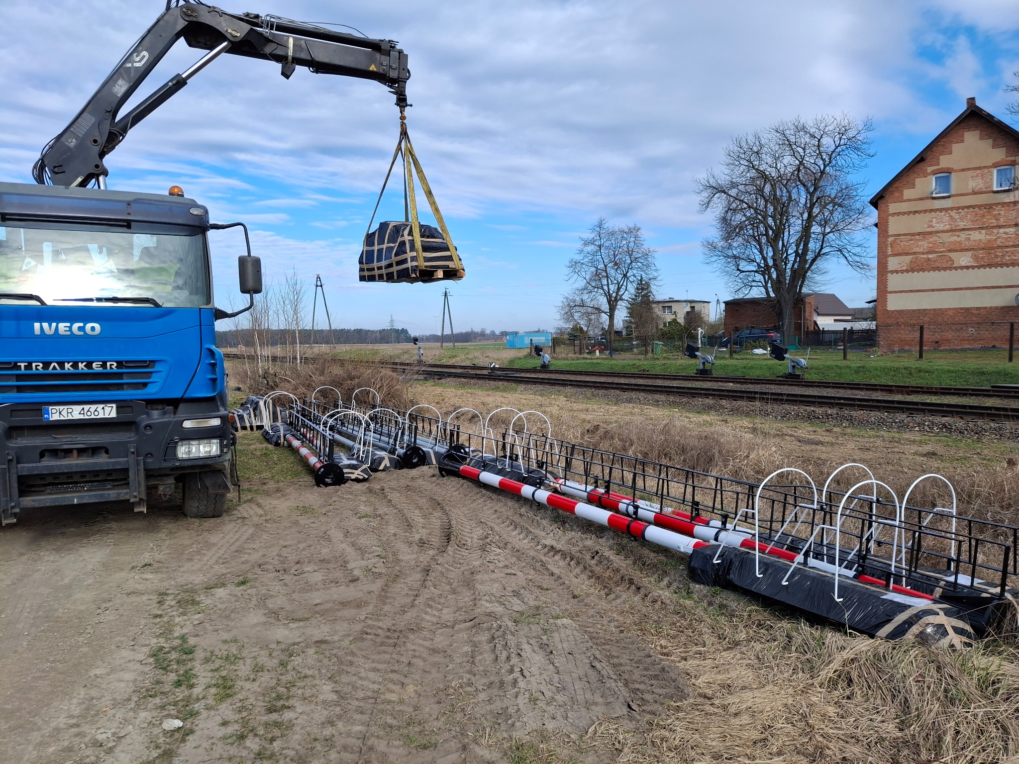 Niebieski samochód ciężarowy z dźwigiem podnosi zapakowany ładunek obok torów kolejowych. Na ziemi leżą elementy infrastruktury kolejowej.