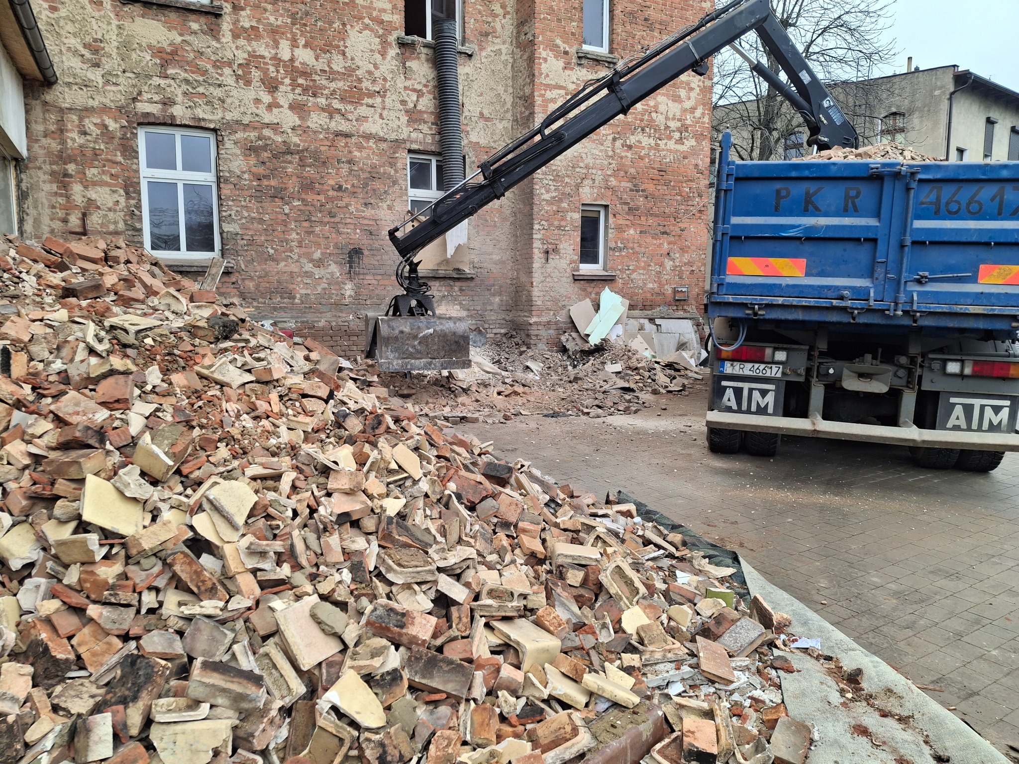 Kopuła gruzu ceglanego przed budynkiem z cegły, załadunek na ciężarówkę z dźwigiem. Widoczne fragmenty okien i instalacji. Prace rozbiórkowe w toku.