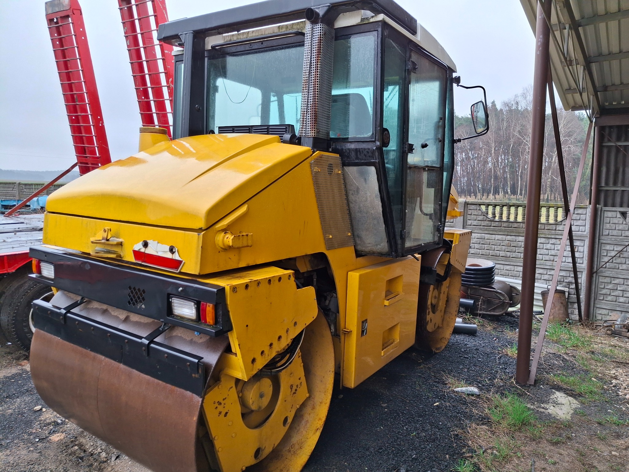 Żółty walec drogowy marki Bomag z widocznymi śladami użytkowania, zaparkowany na utwardzonym terenie z elementami zieleni. Widoczne elementy konstrukcyjne walca i kabina operatora.