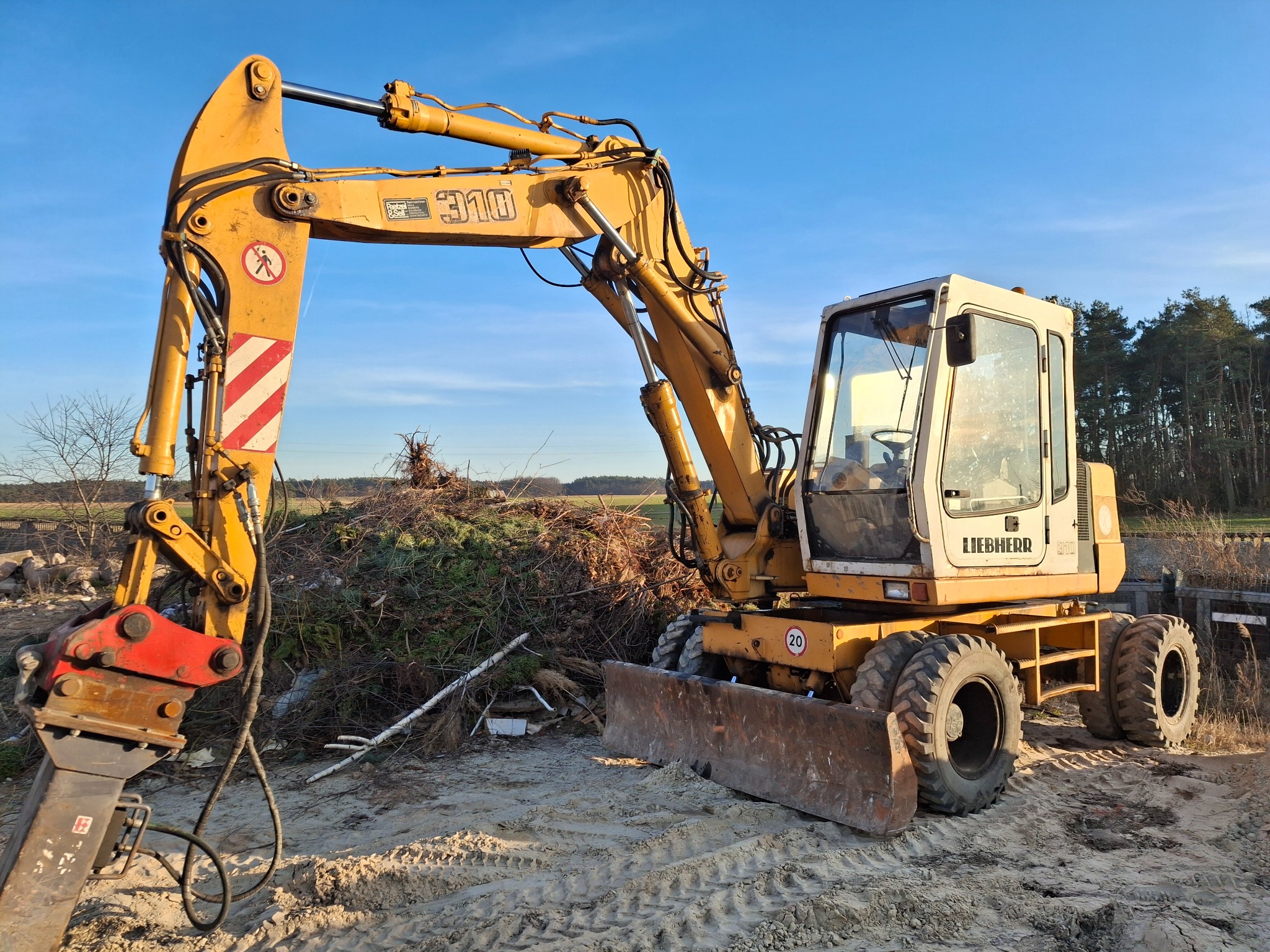 Żółta koparka Liebherr 310 z młotem hydraulicznym na piaszczystym terenie, w tle sterta gałęzi i zieleni, błękitne niebo. Widoczne ślady opon.