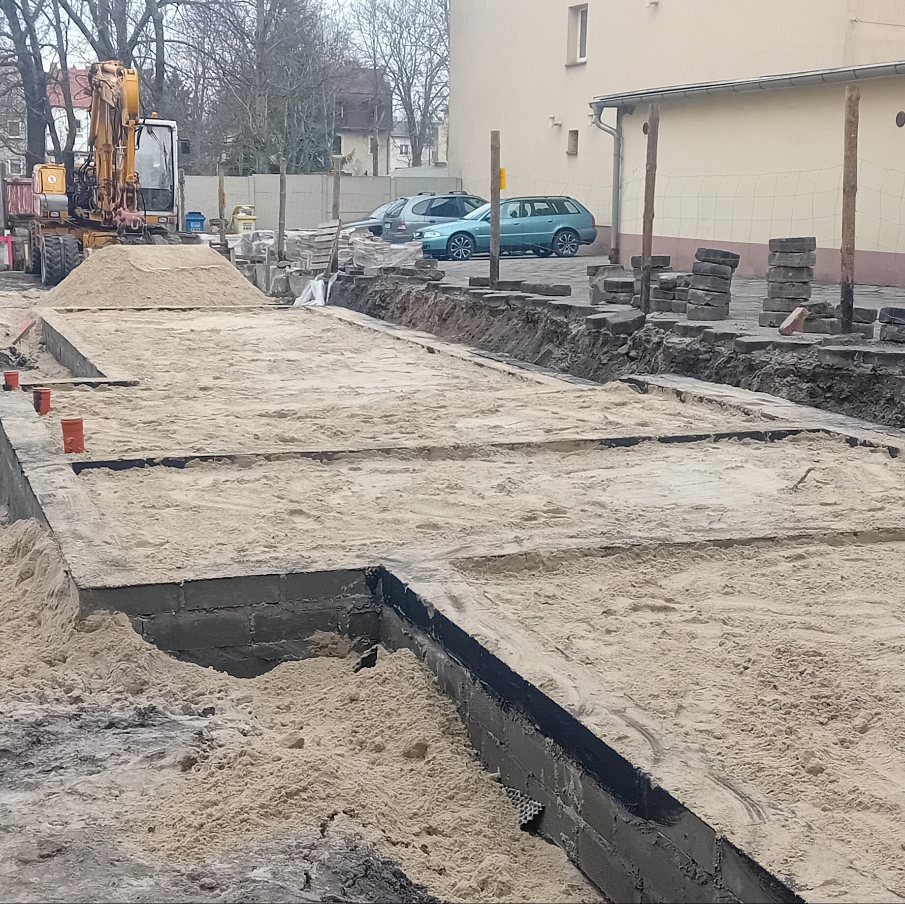 Przygotowanie podłoża z piasku i fundamentów pod kostkę brukową. Na drugim planie koparka i zaparkowane samochody. Prace ziemne w toku.