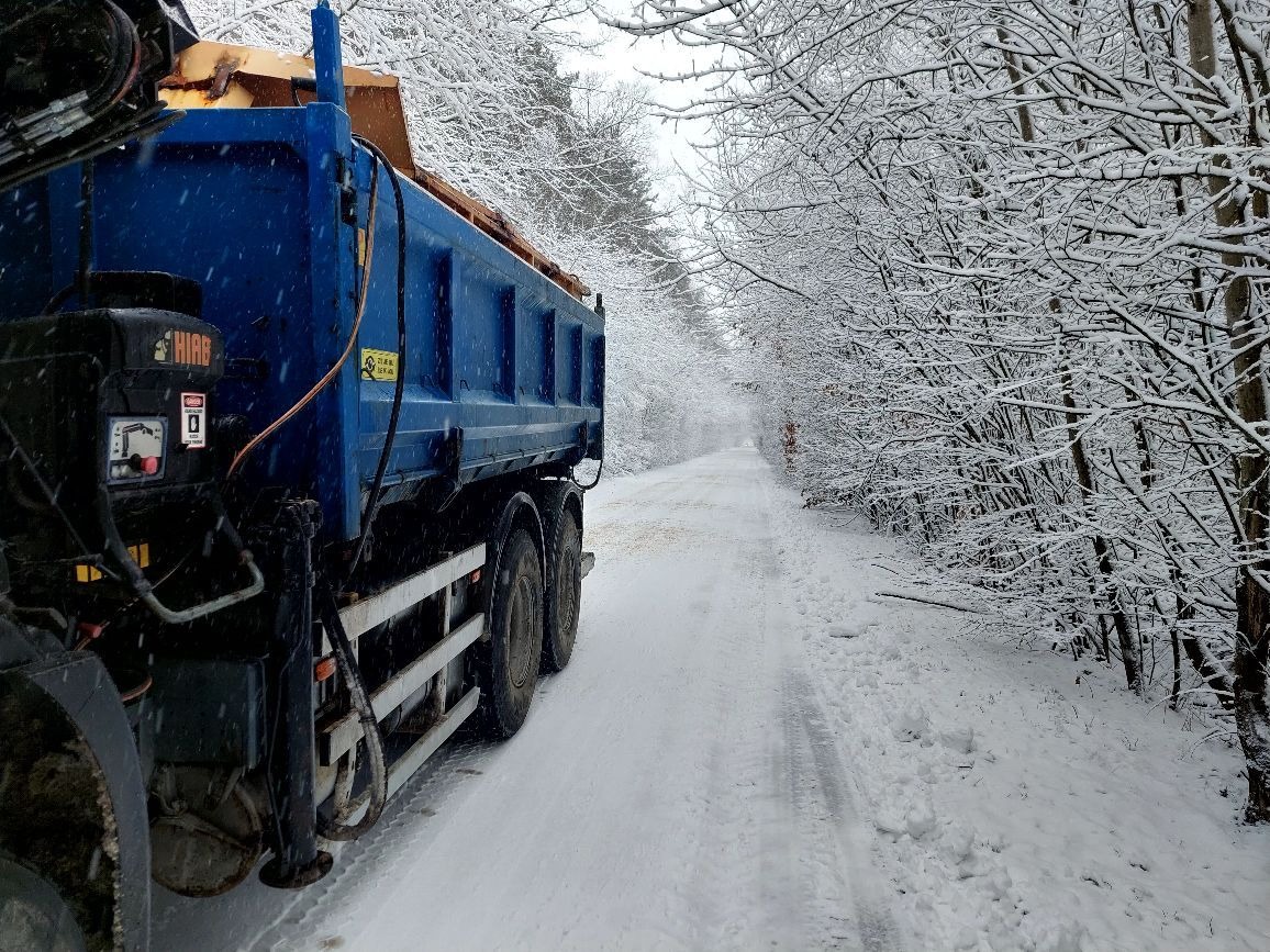 Ciężarówka wywrotka w zimowej scenerii, śnieżna droga leśna. Niebieska skrzynia ładunkowa, widoczne koła i fragment kabiny. Ośnieżone drzewa w tle.