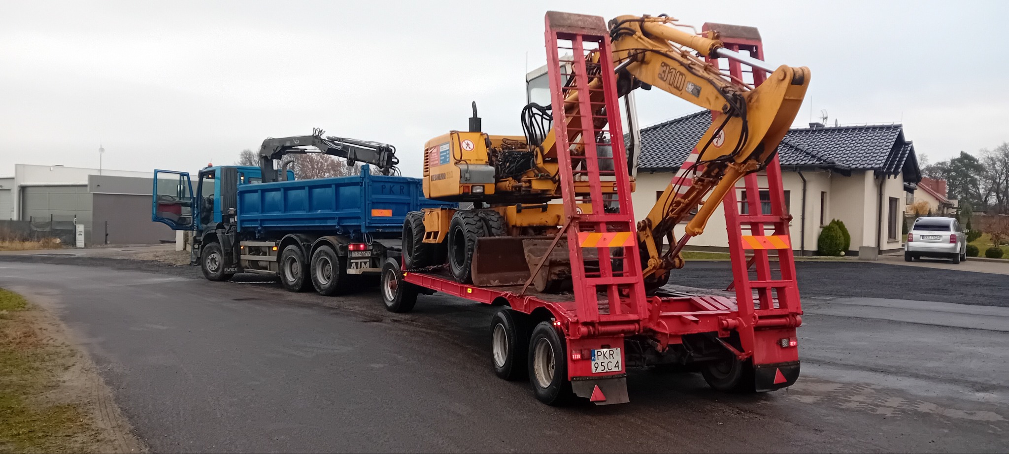 Ciężarówka z dźwigiem holuje na lawecie żółtą koparkę kołową, widok z tyłu, na tle budynki.