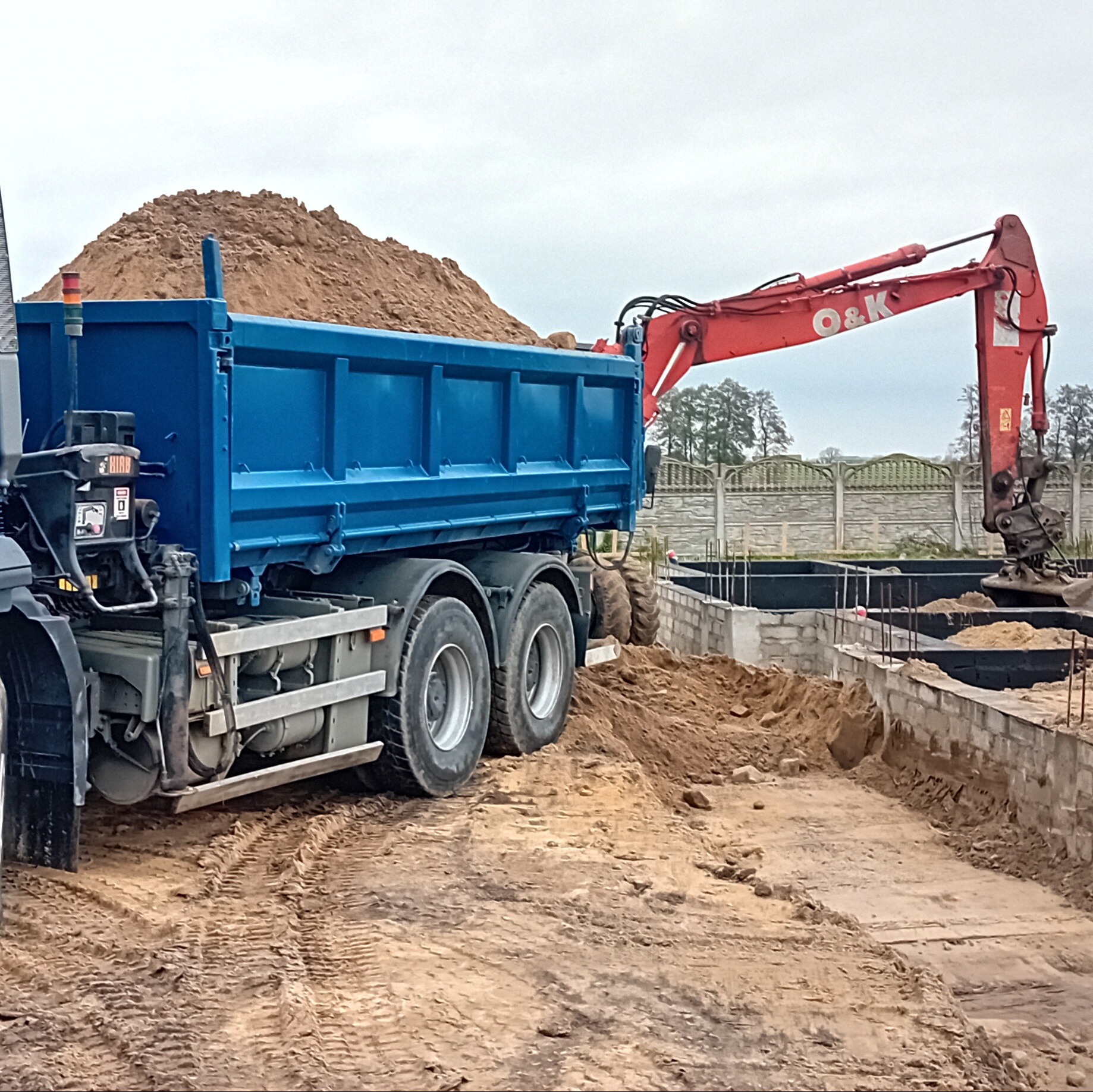 Ciężarówka z piaskiem i koparka O&K przy fundamentach budynku. Roboty ziemne w toku, widoczne ślady opon na placu budowy i ogrodzenie w tle.
