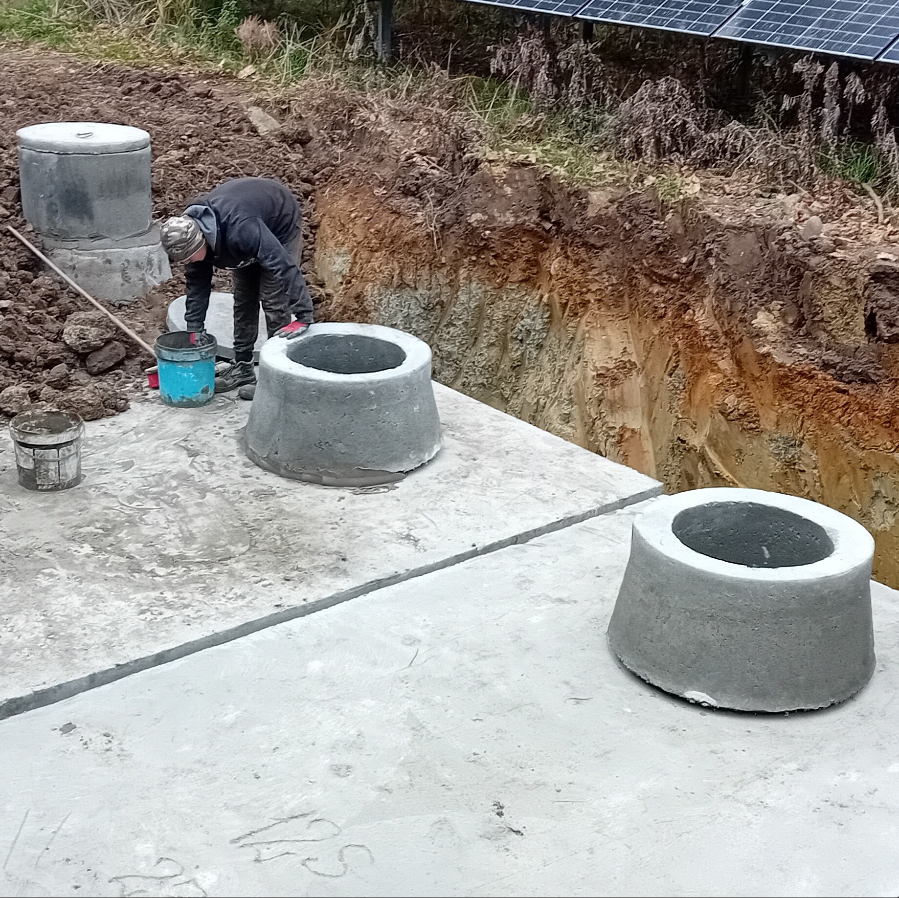Montaż betonowych kręgów studni na placu budowy. Widoczny wykop, częściowo ułożone płyty i osoba w trakcie pracy. W tle panele słoneczne.