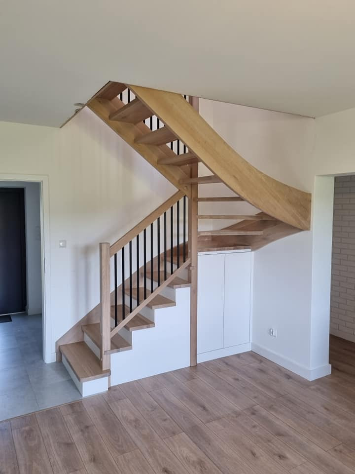 Drewniane schody z czarnymi balustradami i zabudową szafki pod schodami w jasnym wnętrzu z panelami podłogowymi.