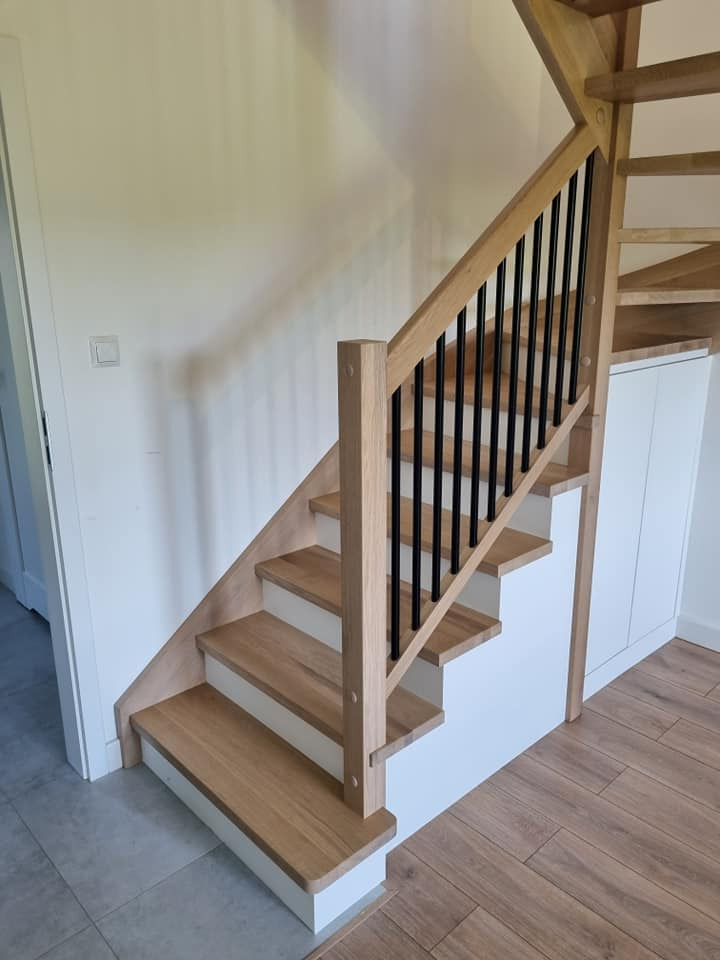 Drewniane schody z czarnymi balustradami i białymi podstopnicami, prowadzące do góry, z zabudową szafkową pod biegiem schodów.