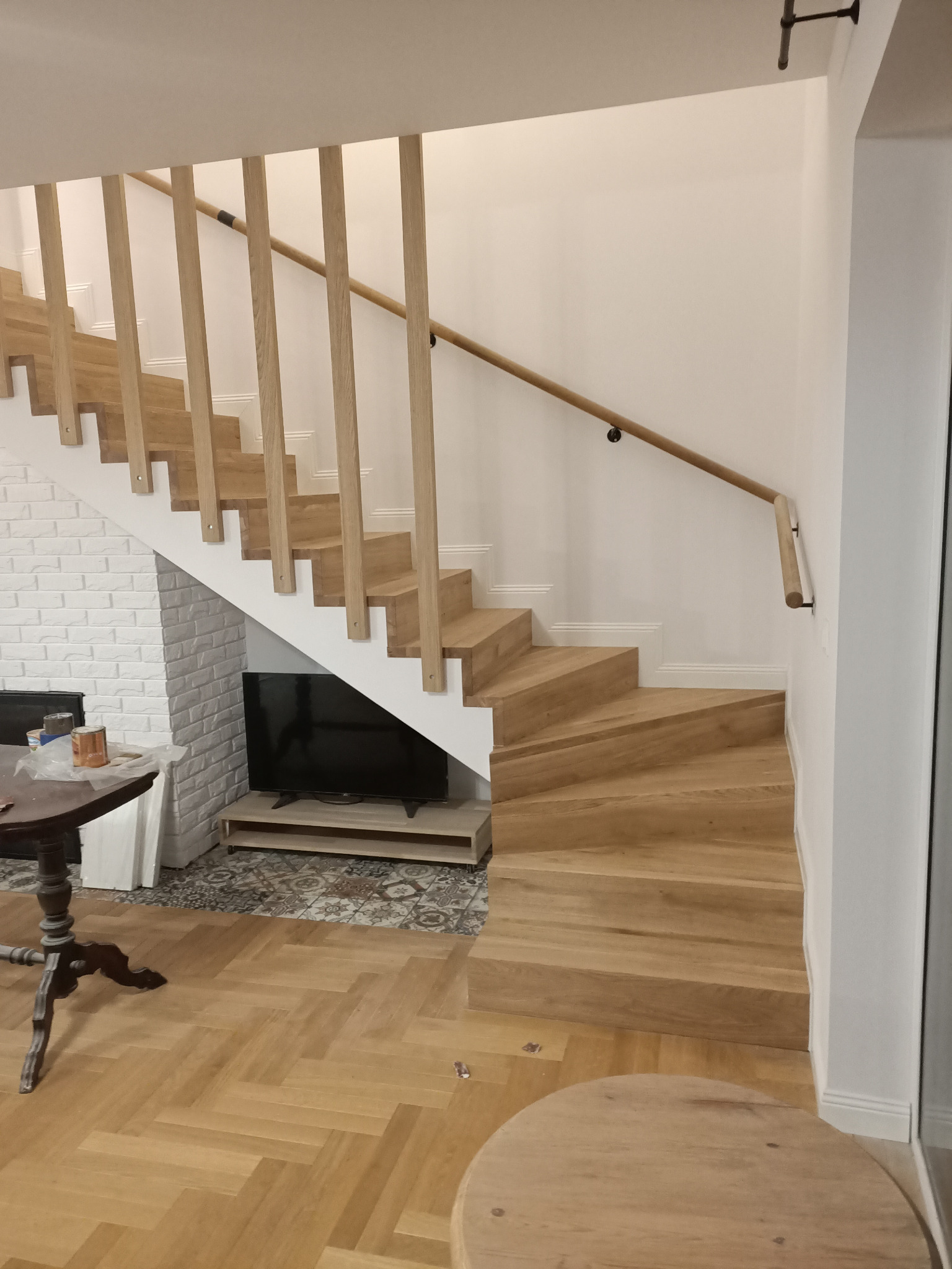 Nowoczesne schody drewniane z jasnego drewna z minimalistyczną balustradą w jasnym wnętrzu z parkietem w jodełkę i ceglaną ścianą w tle.