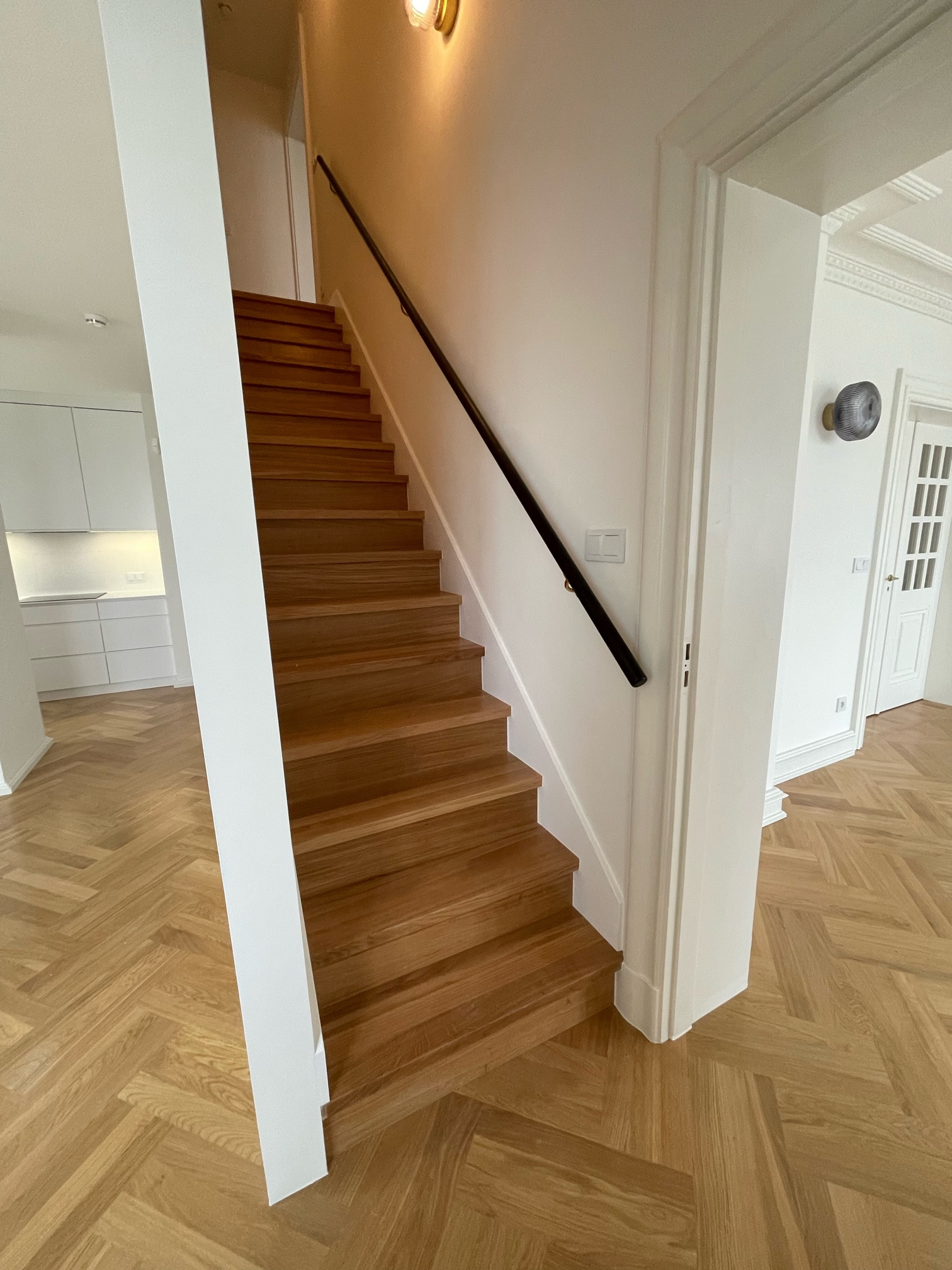 Jasne, drewniane schody z czarną poręczą w nowoczesnym domu, parkiet ułożony w jodełkę, widok na białą kuchnię w tle.