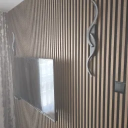 Telewizor zawieszony na ścianie wykończonej panelami pionowymi z drewna, oświetlony dwoma nowoczesnymi lampami ściennymi w kształcie fal.