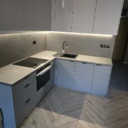 Nowoczesna kuchnia w odcieniach szarości z drewnopodobną podłogą ułożoną w jodełkę, z podświetleniem LED pod szafkami wiszącymi i niedokończonym montażem lampy nad blatem.