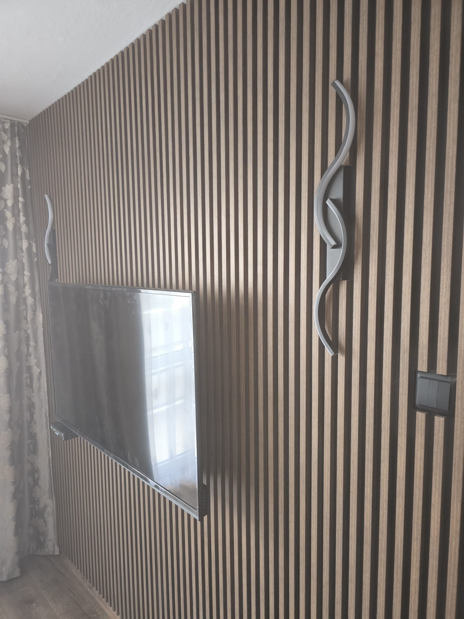 Telewizor zawieszony na ścianie wykończonej panelami pionowymi z drewna, oświetlony dwoma nowoczesnymi lampami ściennymi w kształcie fal.