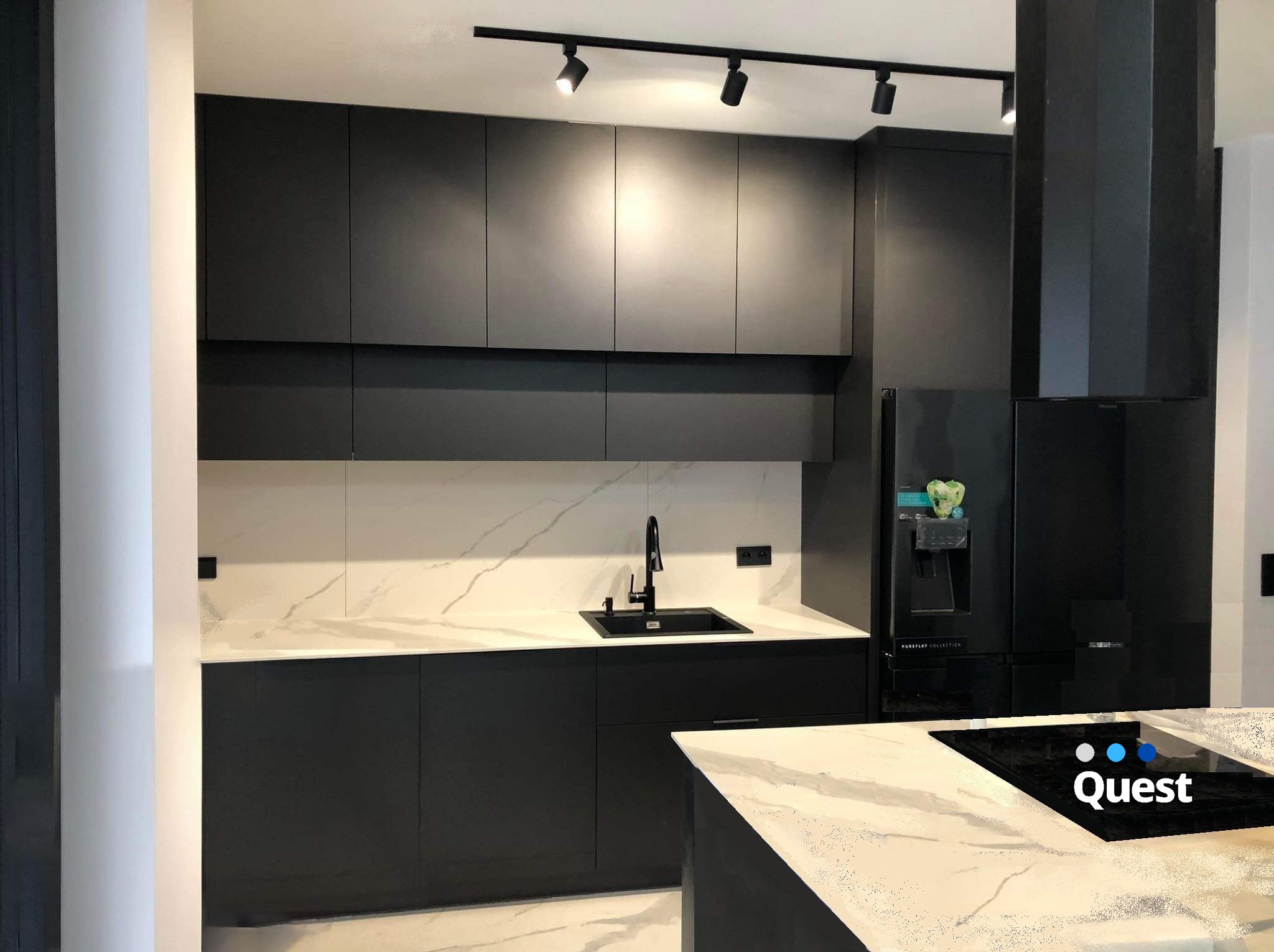 Nowoczesna kuchnia w czerni z białym marmurowym blatem i oświetleniem punktowym, widoczny fragment wyspy kuchennej z płytą indukcyjną.