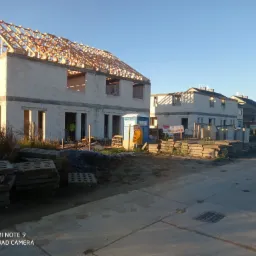 Wczesna faza budowy szeregu domów z pustaków, z drewnianą konstrukcją dachu w pierwszym budynku, przenośna toaleta na placu budowy, widoczne materiały budowlane.