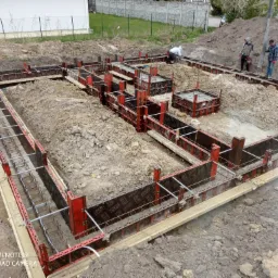 Przygotowanie szalunków i zbrojenia pod fundamenty budynku, widoczne deskowanie z czerwonymi elementami, stalowe pręty zbrojeniowe, ziemia wokół wykopu i pracujący robotnicy w tle.