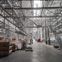 INWEST-REM MONTAŻ SPÓŁKA Z OGRANICZONĄ ODPOWIEDZIALNOŚCIĄ - Wnętrze hali w trakcie budowy, widoczne konstrukcje stalowe, instalacje pod sufitem, maszyny budowlane w oddali oraz palety i materiały budowlane na pierwszym planie.