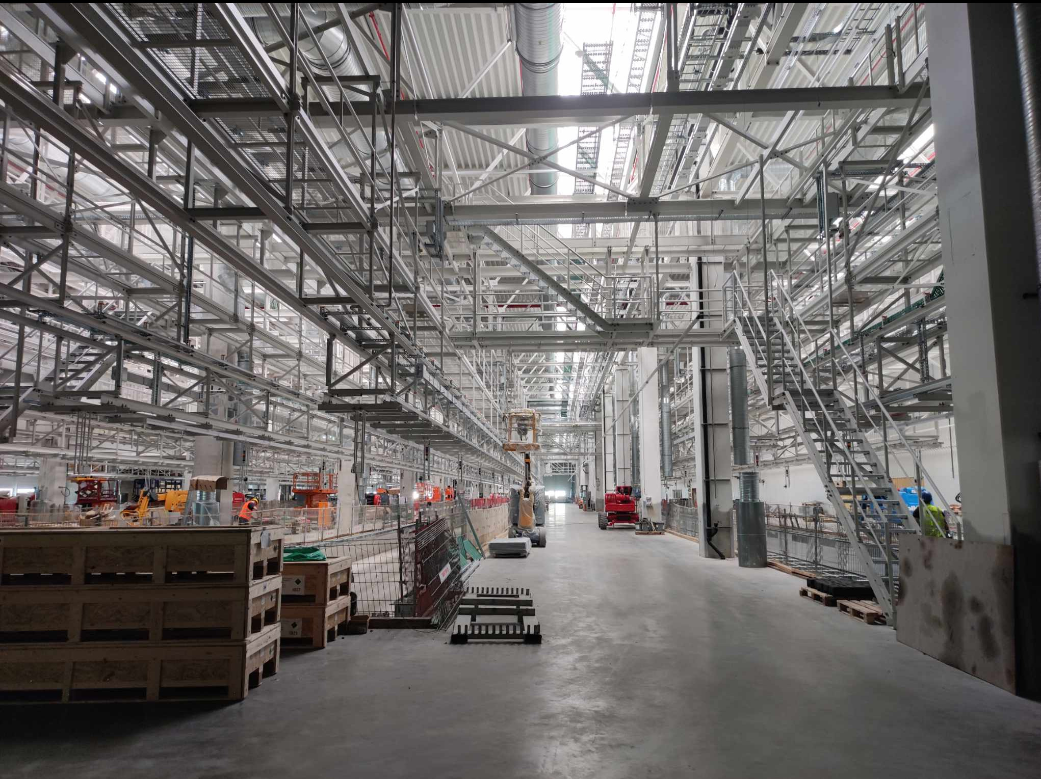 Wnętrze hali w trakcie budowy, widoczne konstrukcje stalowe, instalacje pod sufitem, maszyny budowlane w oddali oraz palety i materiały budowlane na pierwszym planie.