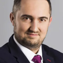 Portret mężczyzny w granatowej marynarce i fioletowym krawacie w kropki, z delikatnym uśmiechem na szarym tle.