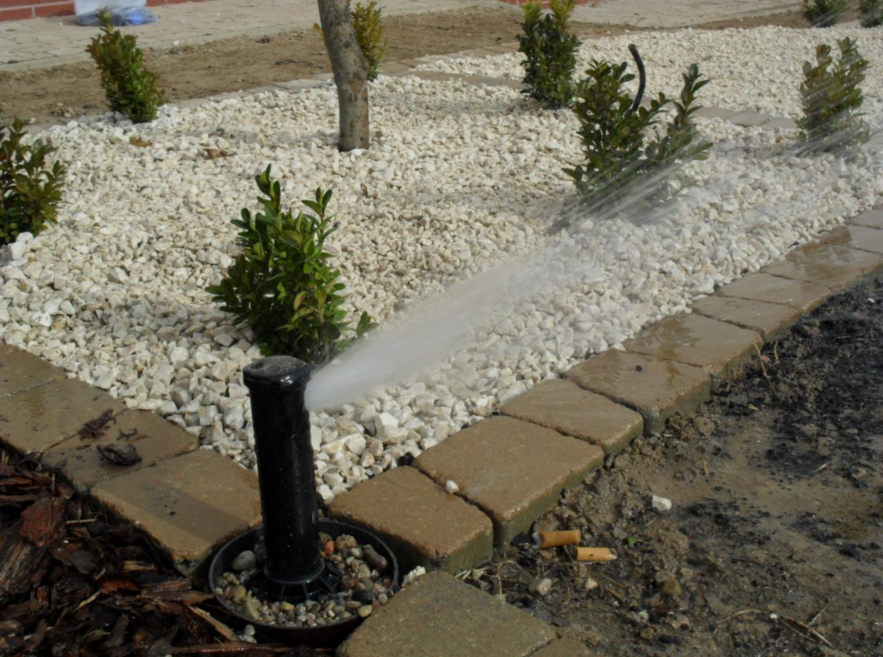 Automatyczny zraszacz nawadnia rabatę z białego grysu, obok obrzeże z kostki brukowej oraz małe krzewy.