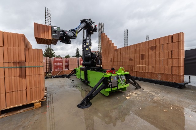 Robot murarski układa cegły na budowie domu z cegły ceramicznej. W tle widoczne zbrojenia i palety z cegłami. Nowoczesna technologia w budownictwie.