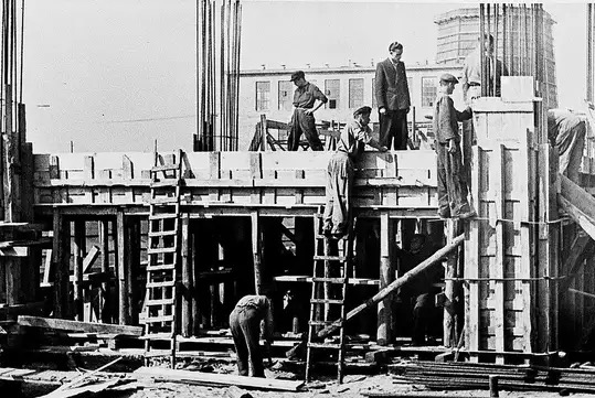 Czarno-białe zdjęcie z budowy fundamentów: robotnicy w trakcie prac zbrojarskich i szalunkowych, widoczne stalowe pręty i drewniane konstrukcje szalunków.