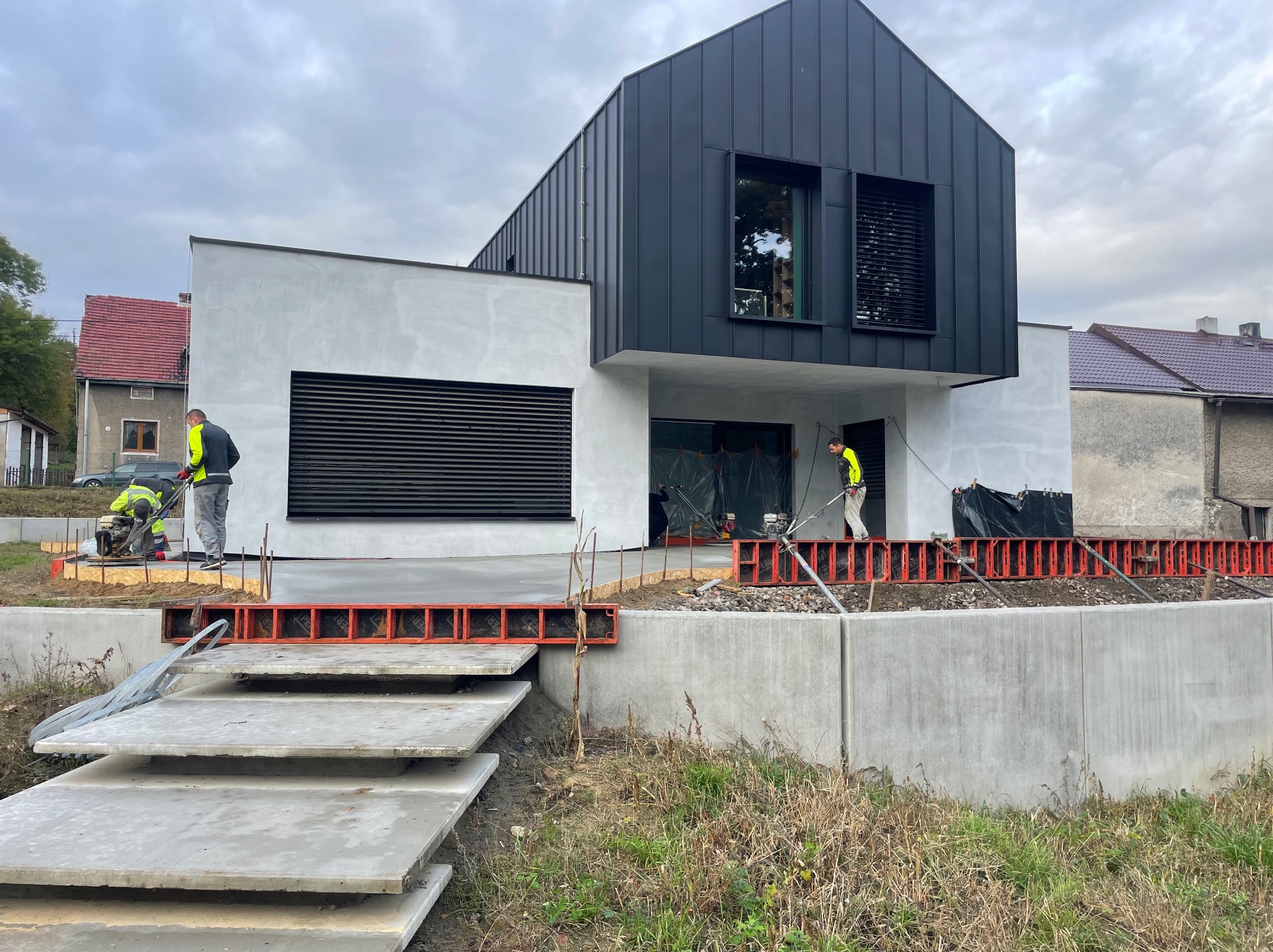 Nowoczesny dom w trakcie budowy: szara elewacja, czarne elementy wykończeniowe. Widoczni pracownicy budowlani i szalunki fundamentowe. Etap wylewania fundamentów.