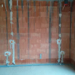 Wnętrze w trakcie instalacji elektrycznej: widoczne przewody w peszlach prowadzone wzdłuż ściany z cegły, zamurowane puszki elektryczne, surowy betonowy strop.