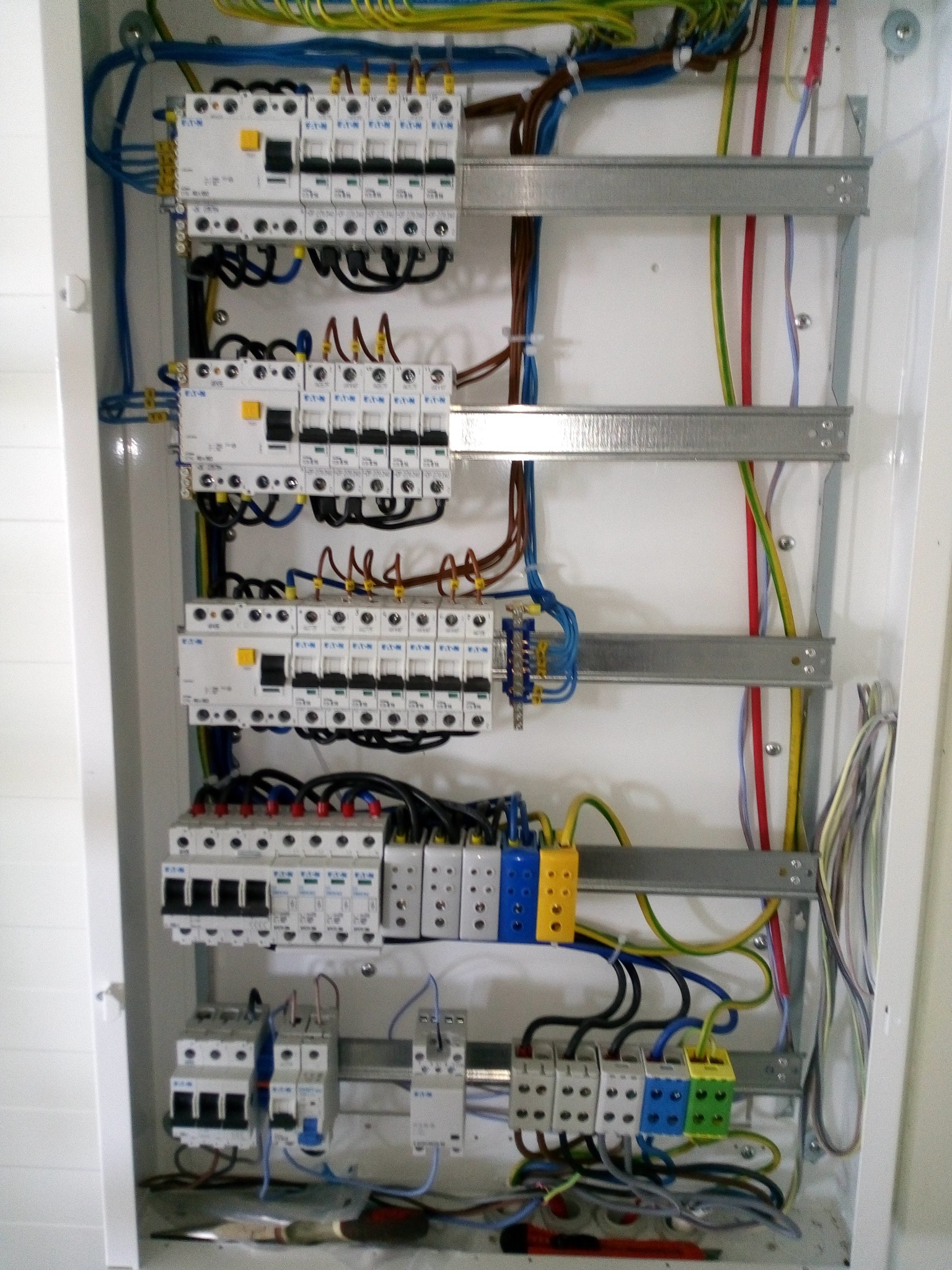 Wnętrze białej skrzynki rozdzielczej z zamontowanymi bezpiecznikami, listwami zaciskowymi, przewodami w różnych kolorach oraz metalowymi szynami montażowymi. Na dnie skrzynki leżą narzędzia.