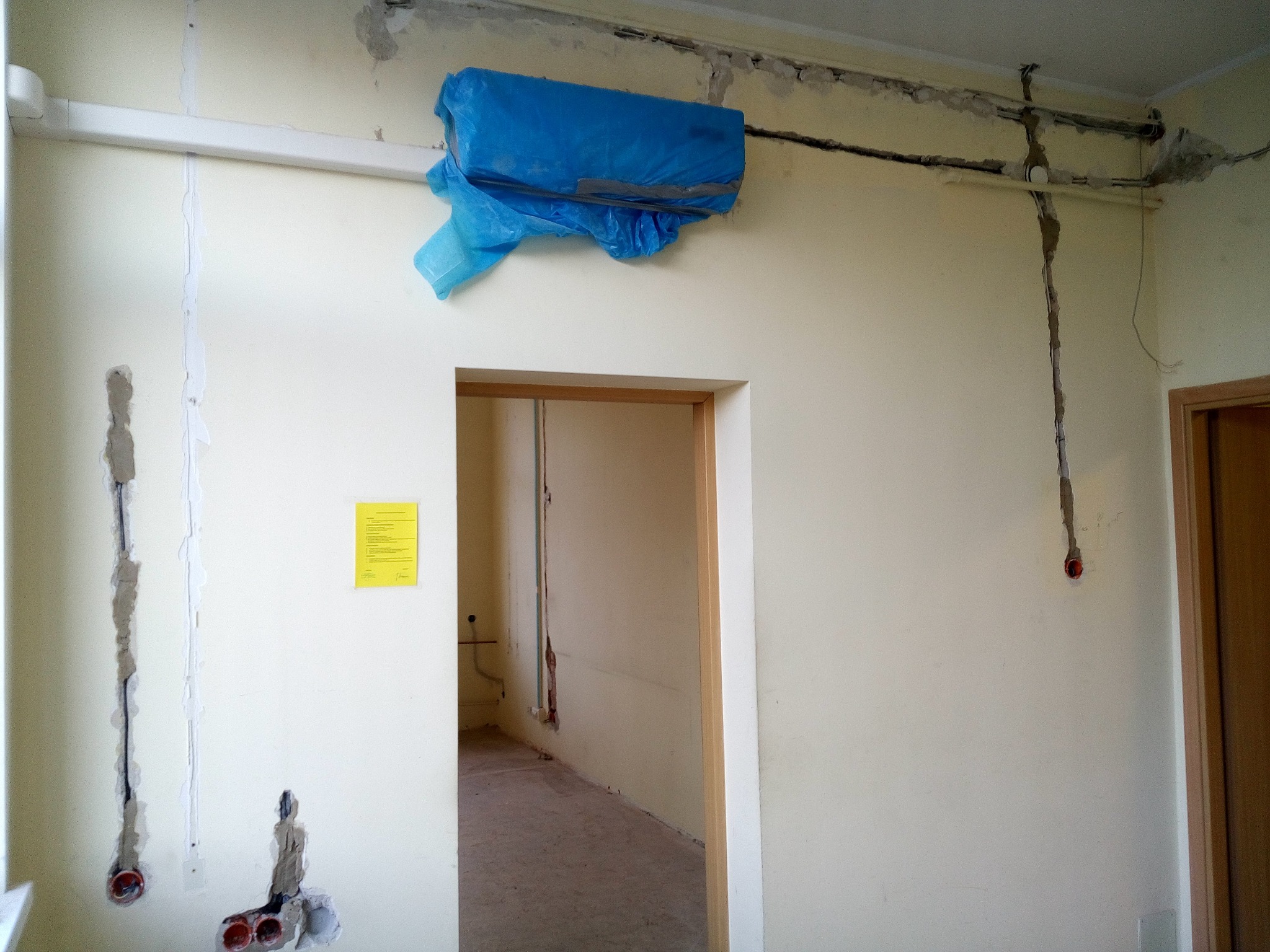 Przygotowanie do rozprowadzenia instalacji elektrycznej wewnątrz budynku: wycięte bruzdy w ścianach, widoczne kable w peszlach, zabezpieczony klimatyzator folią, żółta kartka informacyjna na ścianie.