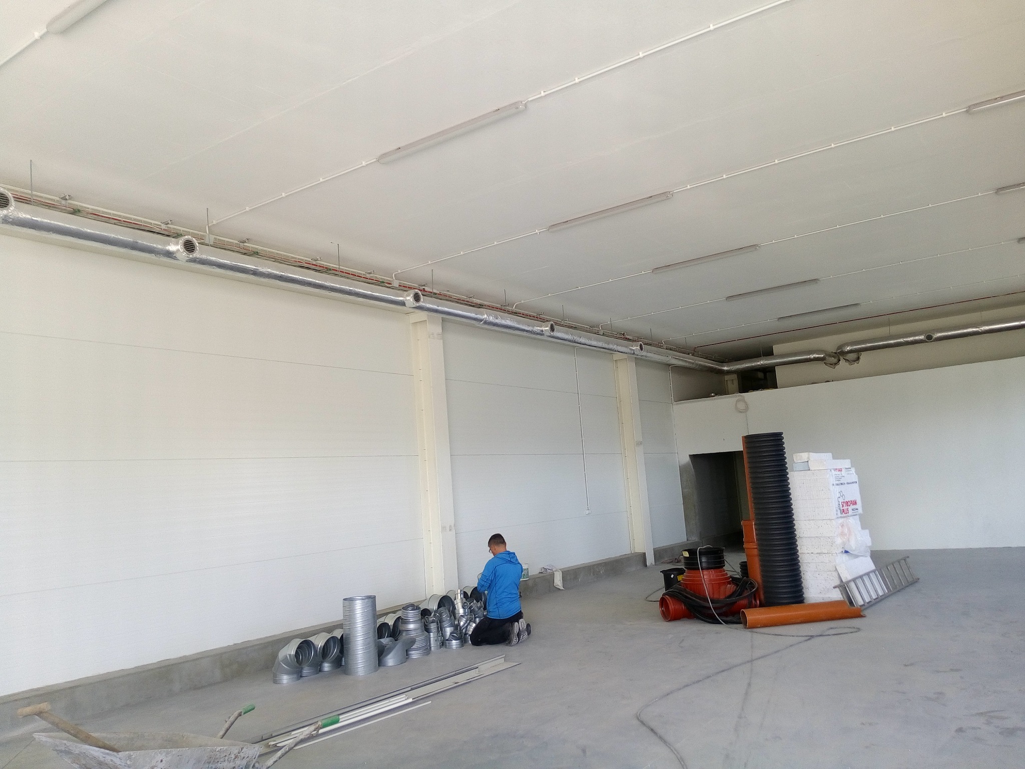 Instalator w niebieskiej bluzie klęczy na podłodze hali, przygotowując elementy wentylacji, widoczne rury wentylacyjne pod sufitem, narzędzia i materiały budowlane w tle.