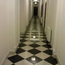 Długi korytarz z biało-czarną, diamentową posadzką z kamienia naturalnego, białe ściany z dekoracyjnym oświetleniem i grzejnikami, ciemne drzwi na końcu korytarza.