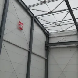 Wnętrze hali przemysłowej w trakcie budowy, widoczne stalowe elementy konstrukcyjne, białe panele ścienne i częściowo odsłonięta betonowa ściana. Czerwono-biała flaga Polski zawieszona na ścianie.