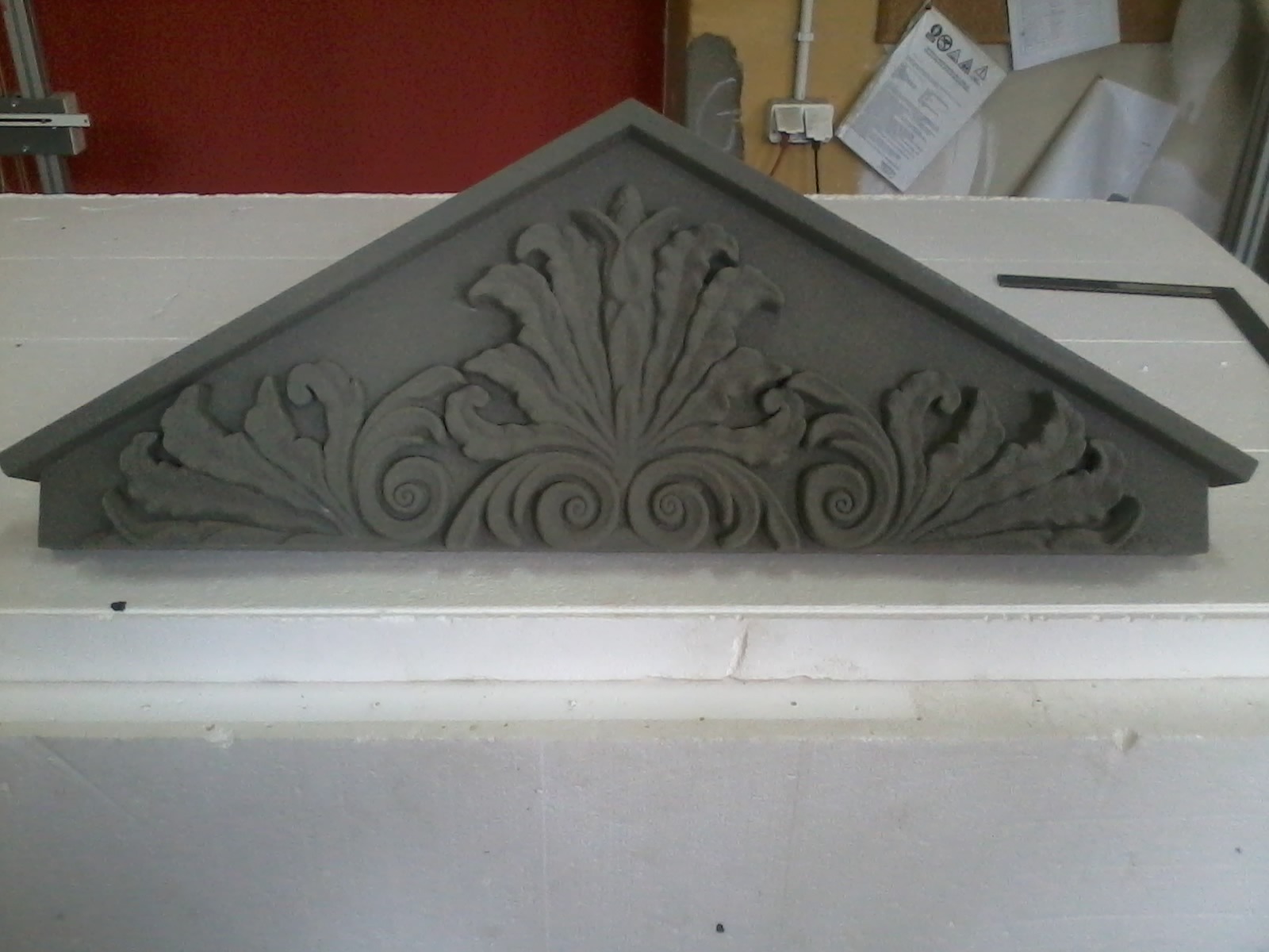 Szary, trójkątny element dekoracyjny z roślinnym ornamentem, leżący na białej powierzchni.