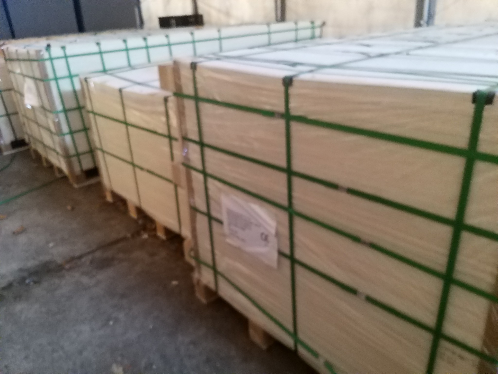 Sztaple paczek materiału izolacyjnego na paletach, owinięte folią i zabezpieczone zielonymi taśmami, widoczne etykiety z oznaczeniami CE.