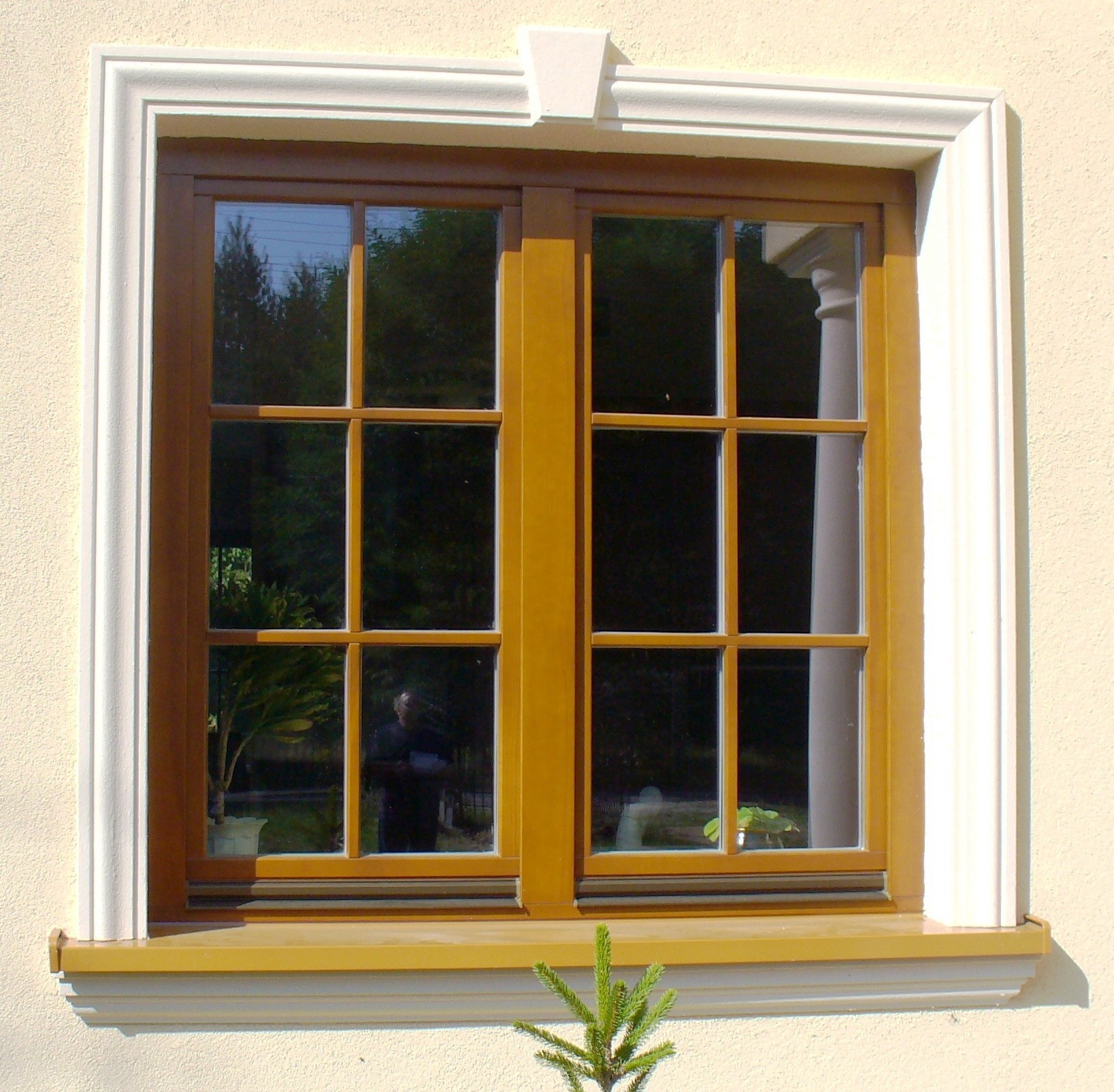 Drewniane okno z białymi ozdobnymi listwami i parapetem, widoczne odbicie osoby w szybie.